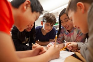 A group of diverse elementary school students collaborate and enjoy working together in a classroom. They are gathered around a desk, holding pencils and smiling as they focus on a shared activity or assignment. The students appear engaged and excited, demonstrating teamwork and learning in a positive environment.