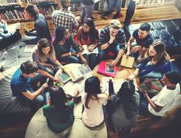 High school students gathered in a collaborative discussion group in a library setting, sitting on couches and around a table, engaged with books, notebooks, and a tablet.