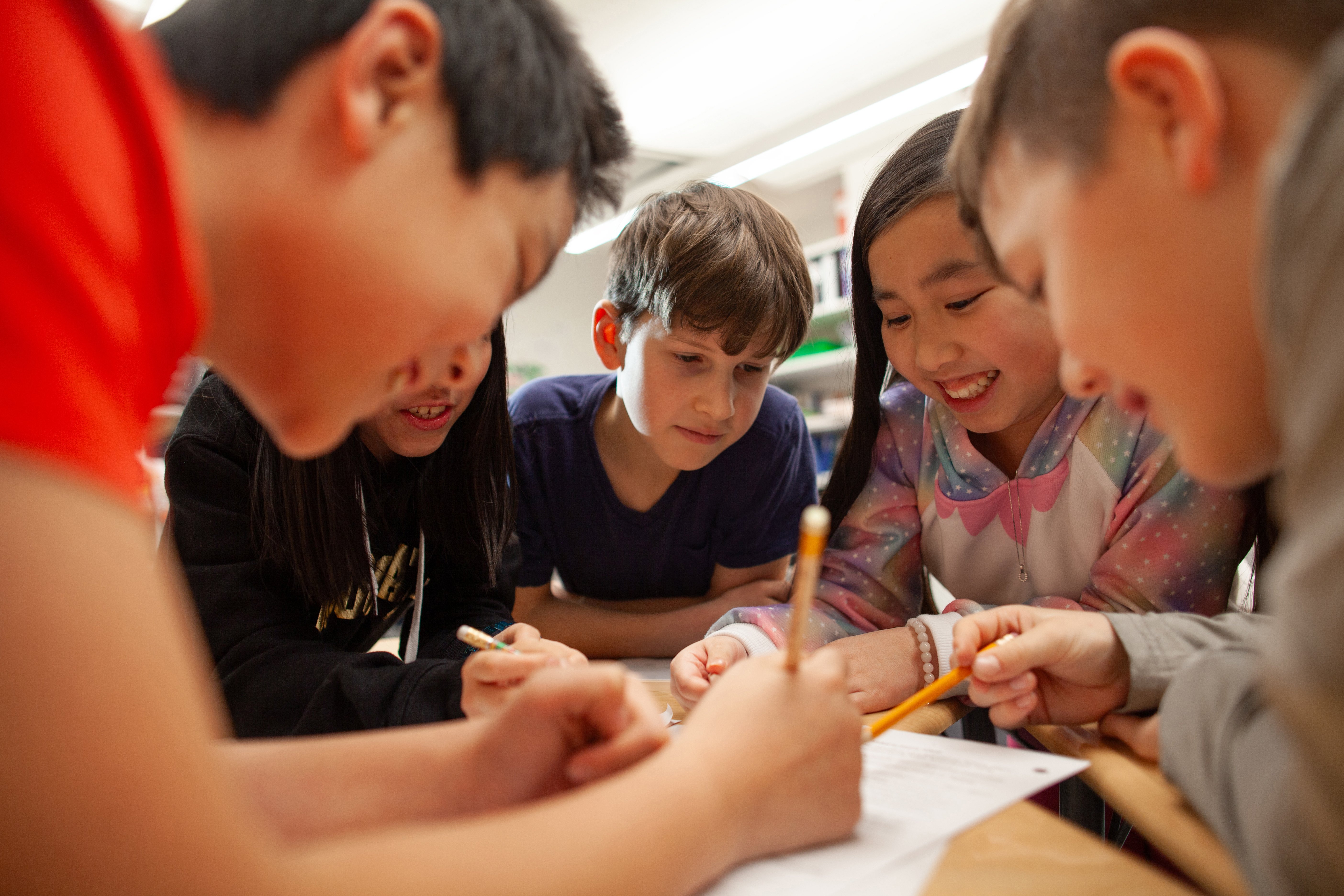  A group of diverse elementary school students collaborate and enjoy working together in a classroom. They are gathered around a desk, holding pencils and smiling as they focus on a shared activity or assignment. The students appear engaged and excited, demonstrating teamwork and learning in a positive environment.